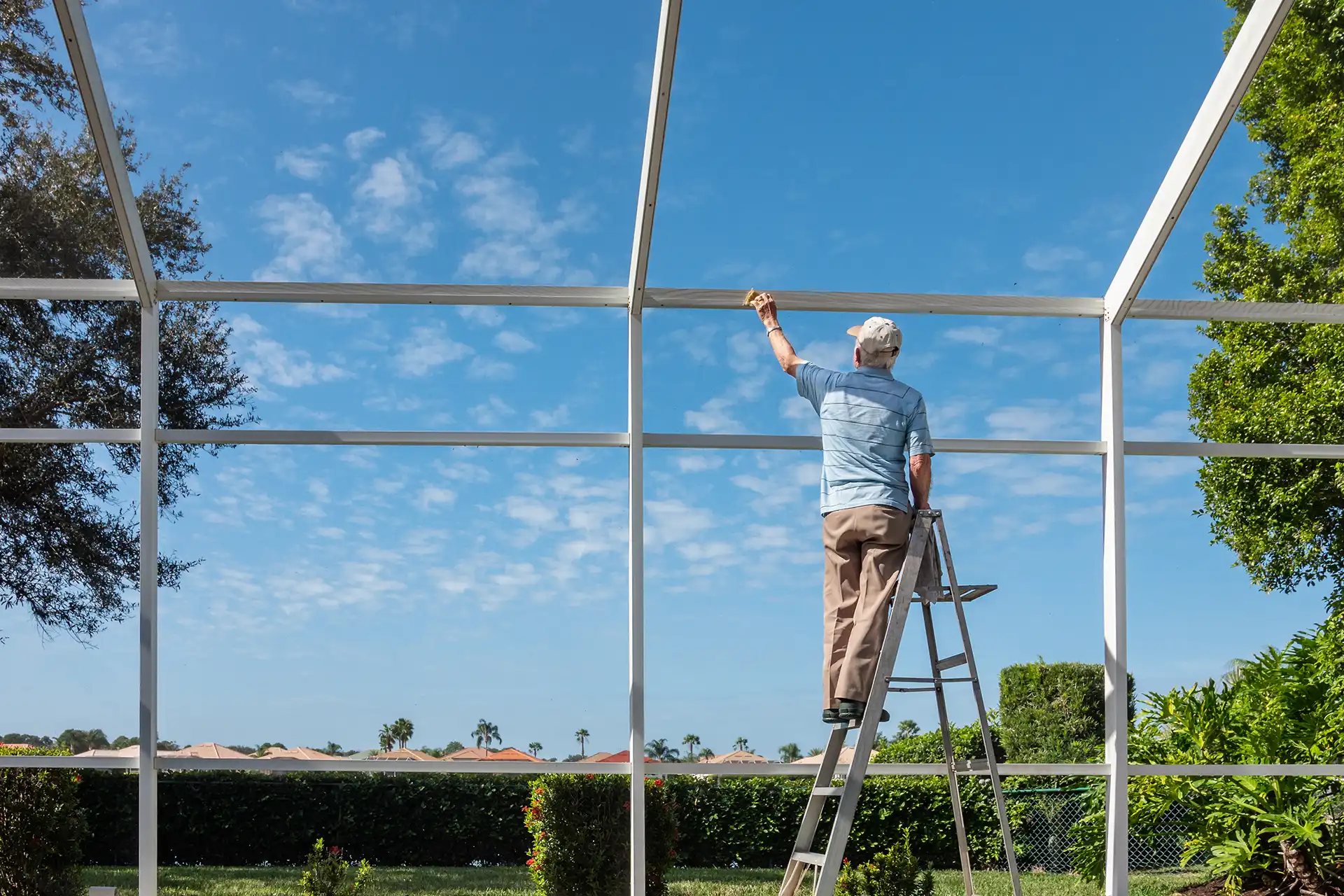 A man on a step ladder performing maintenance on a white screened-in patio enclosure under a blue sky
