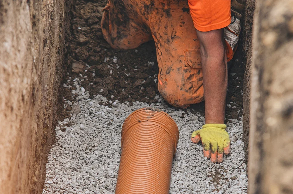 A worker in orange gear placing a corrugated drainage pipe into a gravel-lined trench