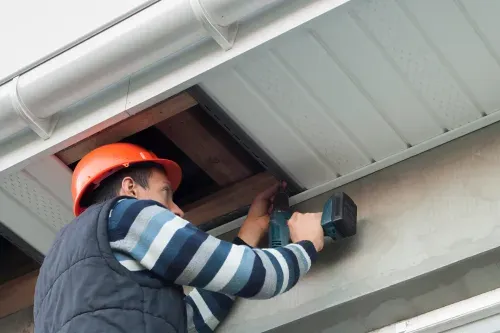 A worker using a power drill to install white soffit panels on a house