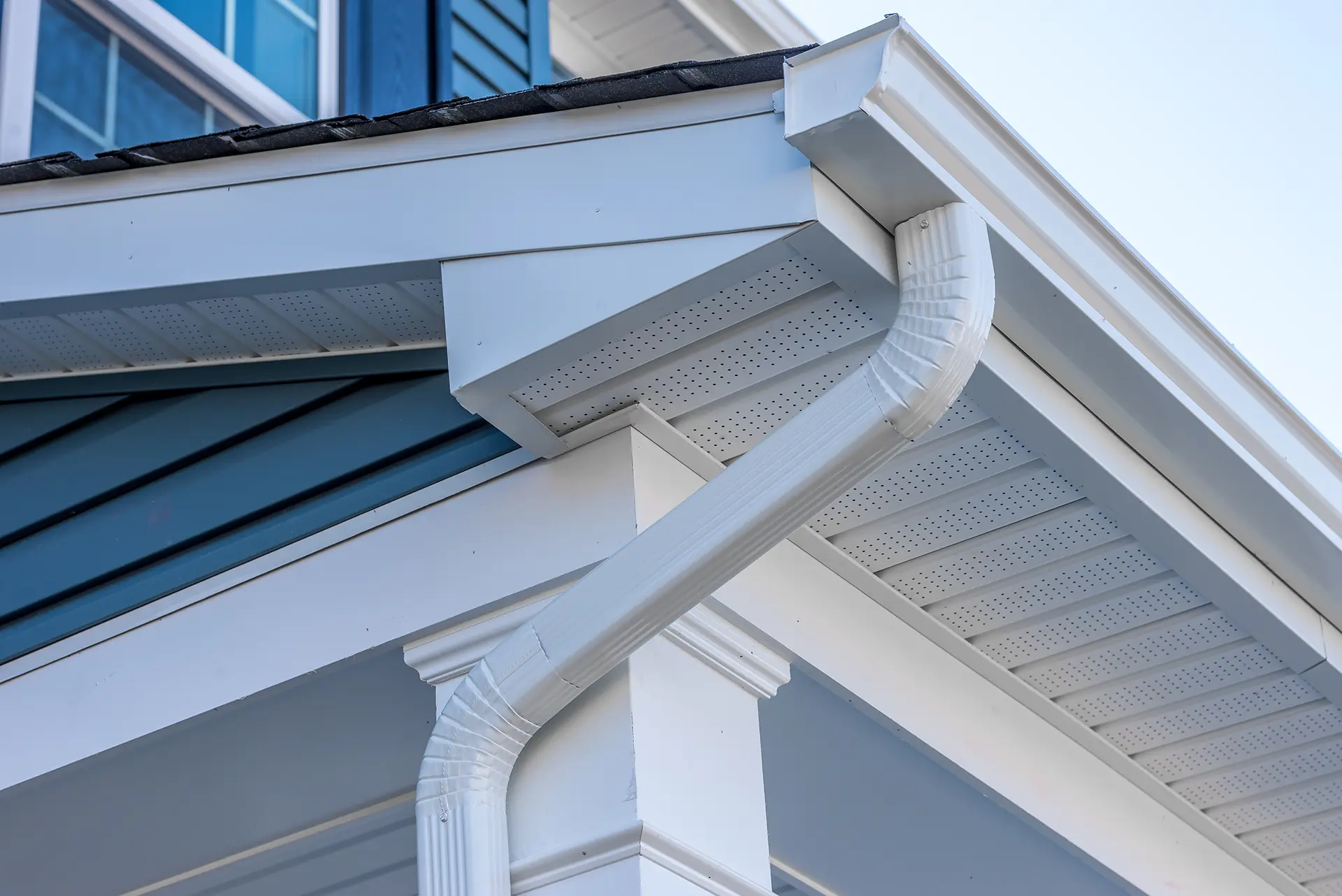 Close-up of white seamless gutters and a downspout installed on a residential house with blue siding