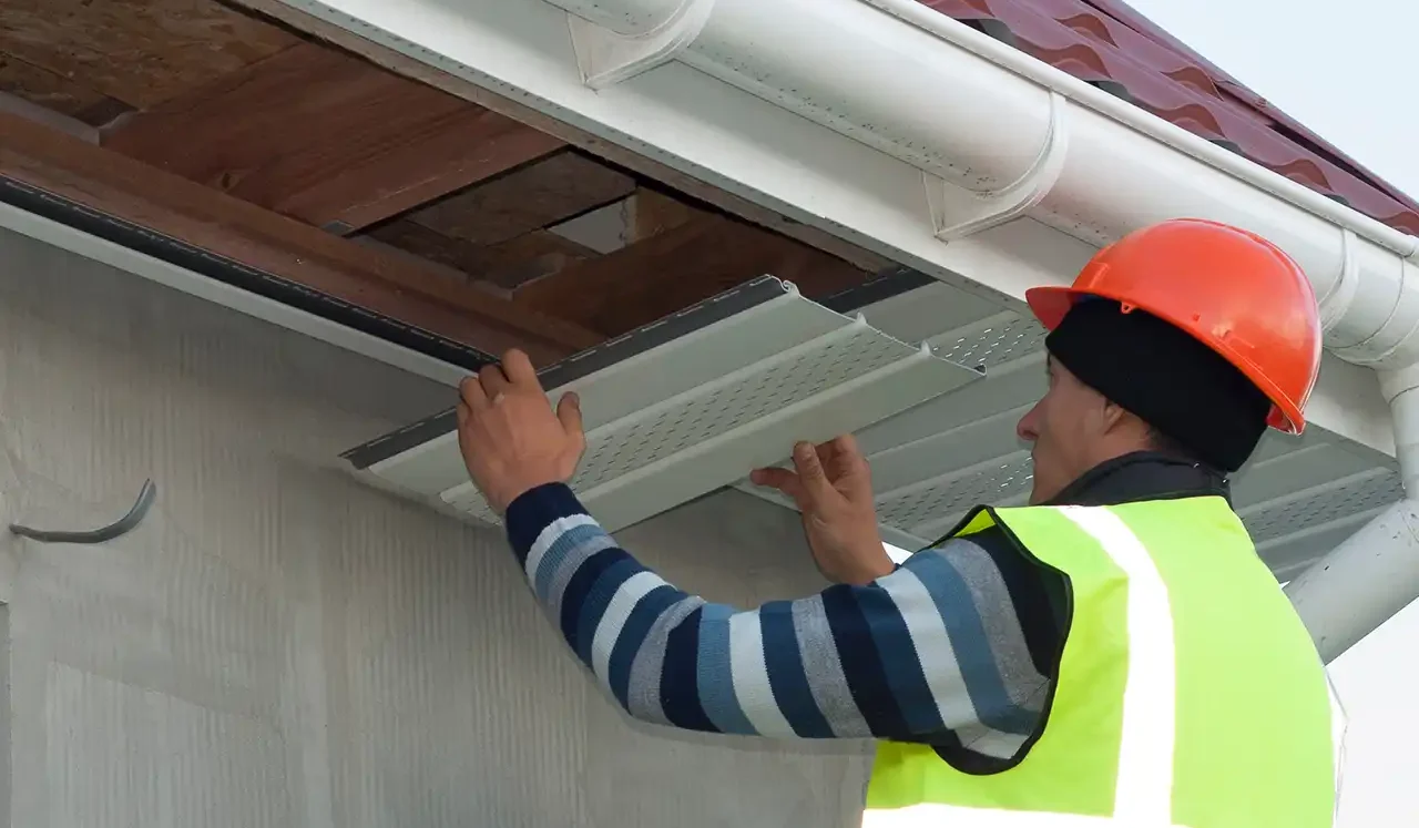 Construction worker in high-visibility gear carefully fitting a white soffit panel into a roof overhang