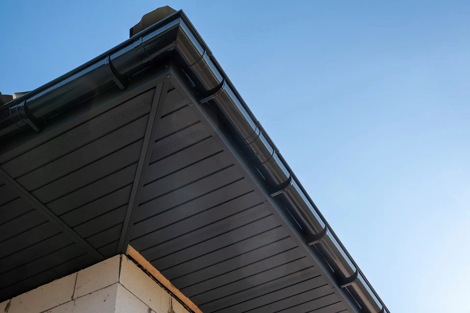 Low-angle view of a modern black gutter system and dark soffit against a clear blue sky