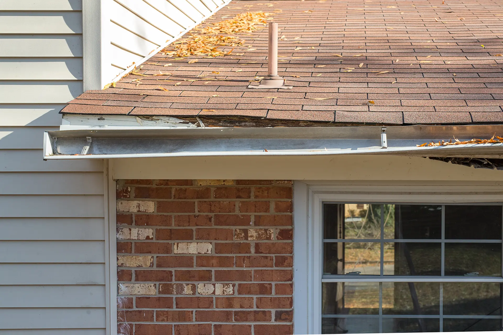 View of old metal gutters filled with dry autumn leaves on a brick and siding house.